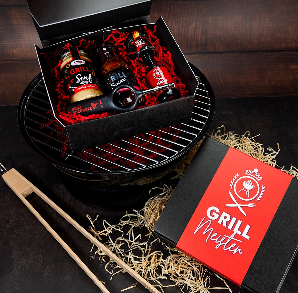 Grillmeister Box auf Grill mit Zange und Stroh im Hintergrund.