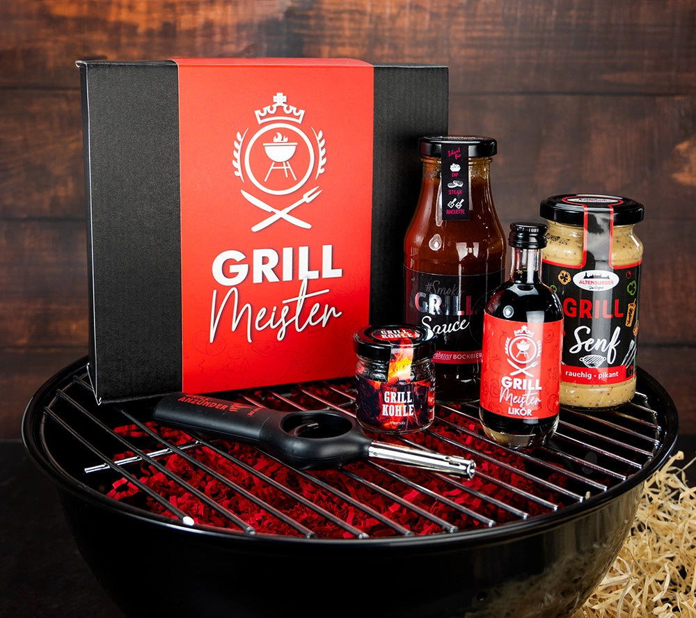 Inhalt Grillmeister Box auf einem Grill. Der Inhalt ist ein Feuerzeug, Grillmeister Sauce, Grillmeister Likör, Grillmeister Senf, Grillmeister Salz "Grill Kohle"